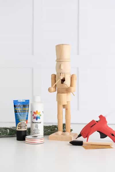 Unpainted wooden nutcracker next to a red hot glue gun, white paint, a foam brush, sandpaper, white and red ribbon, wood filler and faux greenery on a white surface.