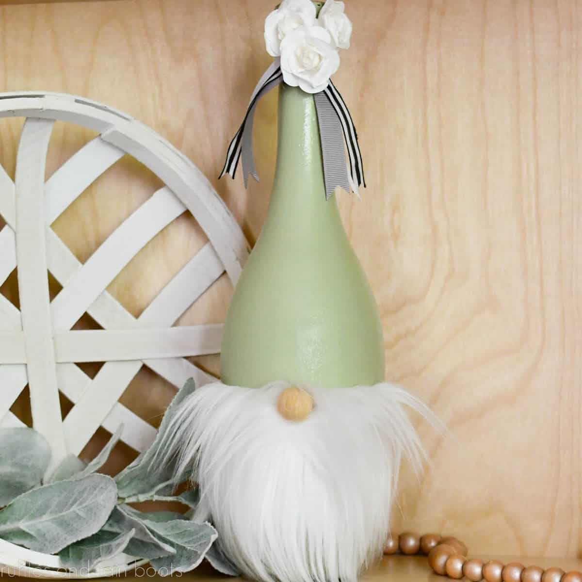 Square close up image of a green painted wine bottle gnome with white beard and flowers on a farmhouse decor background.