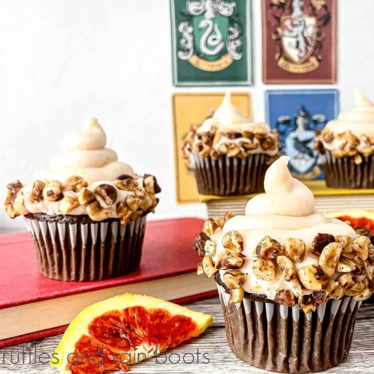 Vertical close up image of a frosted cupcake with hazelnuts, next to 2 slices of a blood orange with 3 cupcakes in the background on top of books and the Hogwarts house emblems on a weathered wood surface.