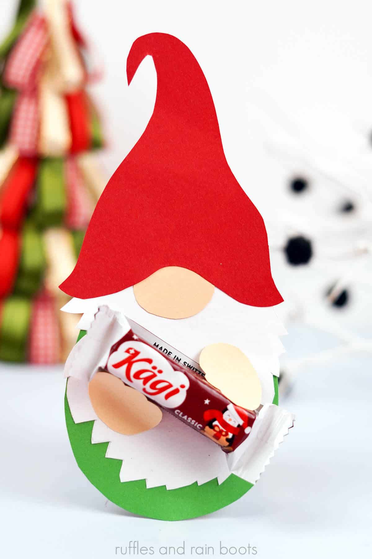 Close up vertical image of a paper gnome with red hat, green body, and hands holding a piece of chocolate.