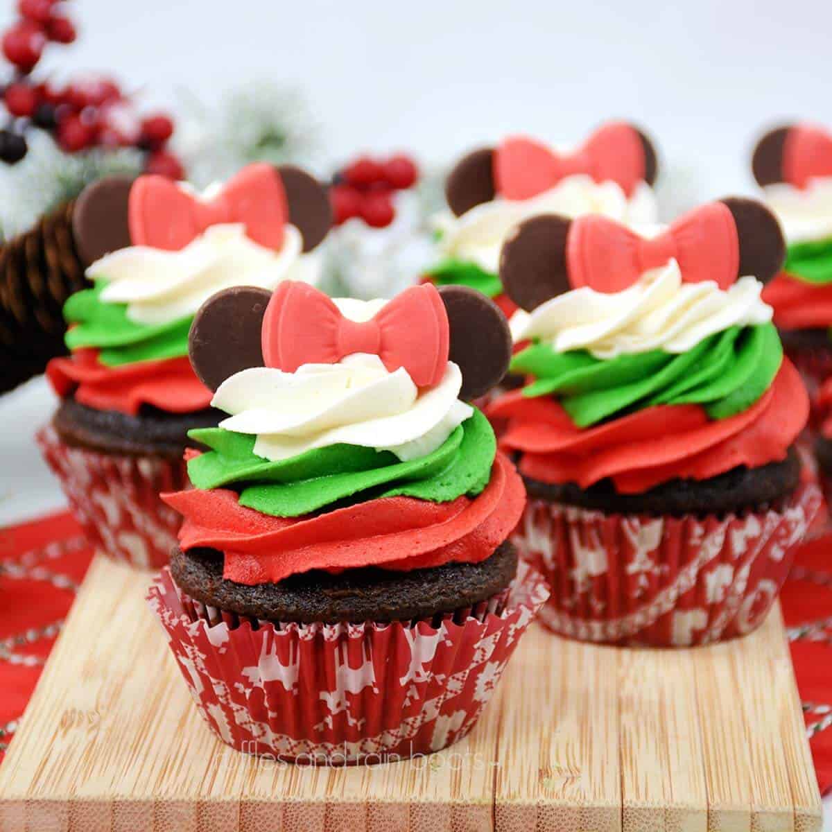 S close up image of 6 frosted Minnie Mouse Christmas cupcakes on a wooden slab on a red and white napkin on a white surface with Christmas greenery in the background.