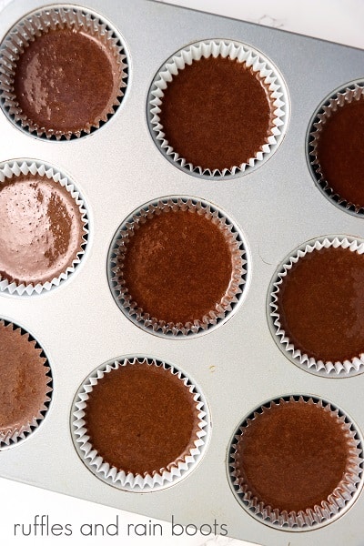 V Overhead image of a metal pan lined with whited cupckae liners with chocolate cupcake batter on a white surface.