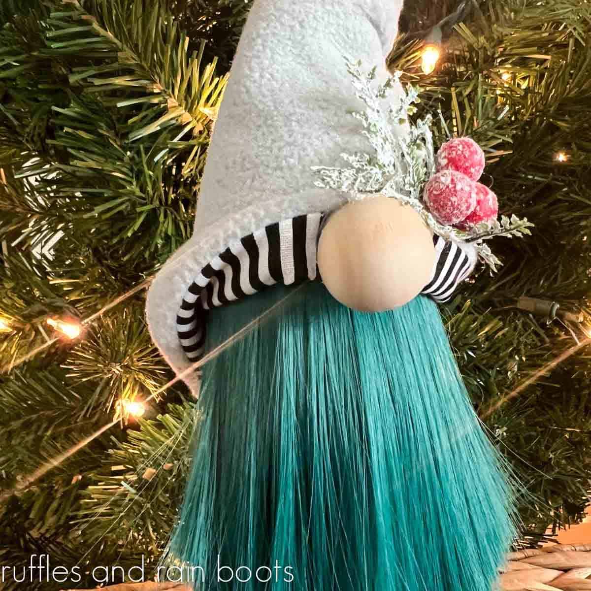 Close up image of a holiday gnome with a green doll hair beard, a reversible gnome hat, and a large wooden nose in front of a Christmas tree.