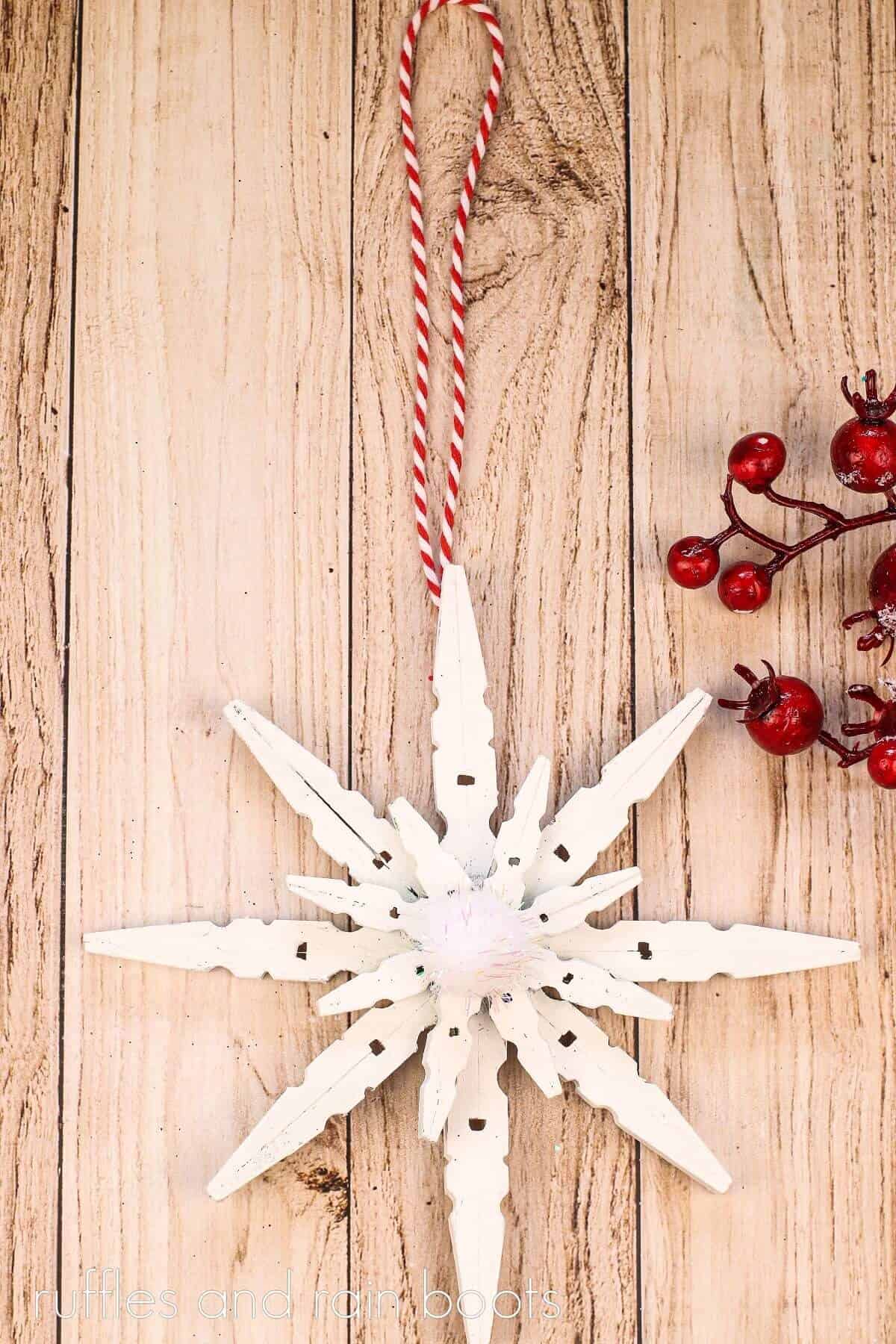 V White layered wooden clothespin snowflake ornament on a wooden surface next to red holiday decor.