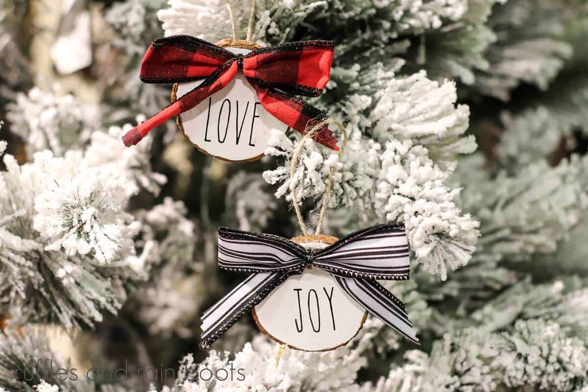 White wood slice ornaments hanging on a white flocked tree, one with a black and white bow and one with a red and black bow with Rae Dunn inspired lettering and twine hanger.