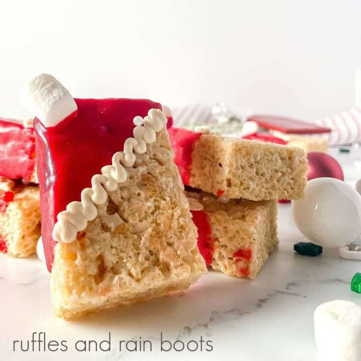 V close up image of a cereal treat dipped in red with a mini marshmallow in the corner on a white surface with holiday decorations around them.