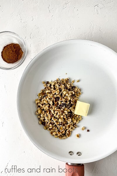 A vertical overhead image of hazelnuts and butter in a large white skillet, next to a small bowl of cinnamon on a white surface.
