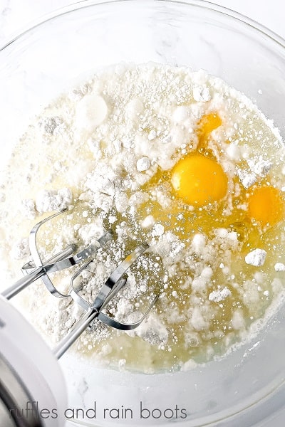 V Overhead image of a large round glass bowl with a cake mix, eggs,water, and oil with an electric hand mixer for the Hedwig Cookies and Cream Cupcakes on a marble surface.