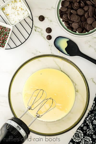 Vertical overhead image of a large round glass bowl and a black hand mixer with sweetened condensed milk with sprinkles in white square bowls, a glass round bowl of candy melts and a black spoon next to black and white fabric on a white marble surface.