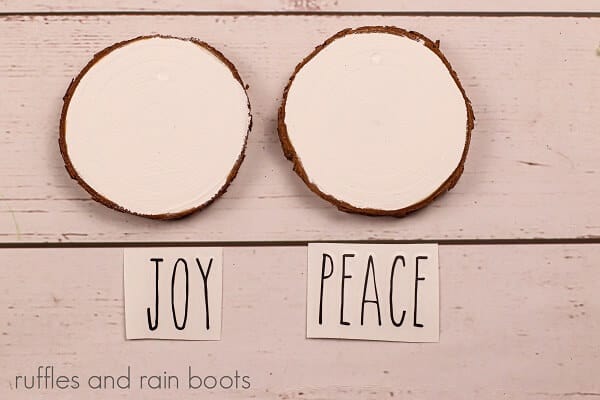 White wooden wood slice ornaments, with decals of black lettering saying joy and one saying peace on a wooden surface.