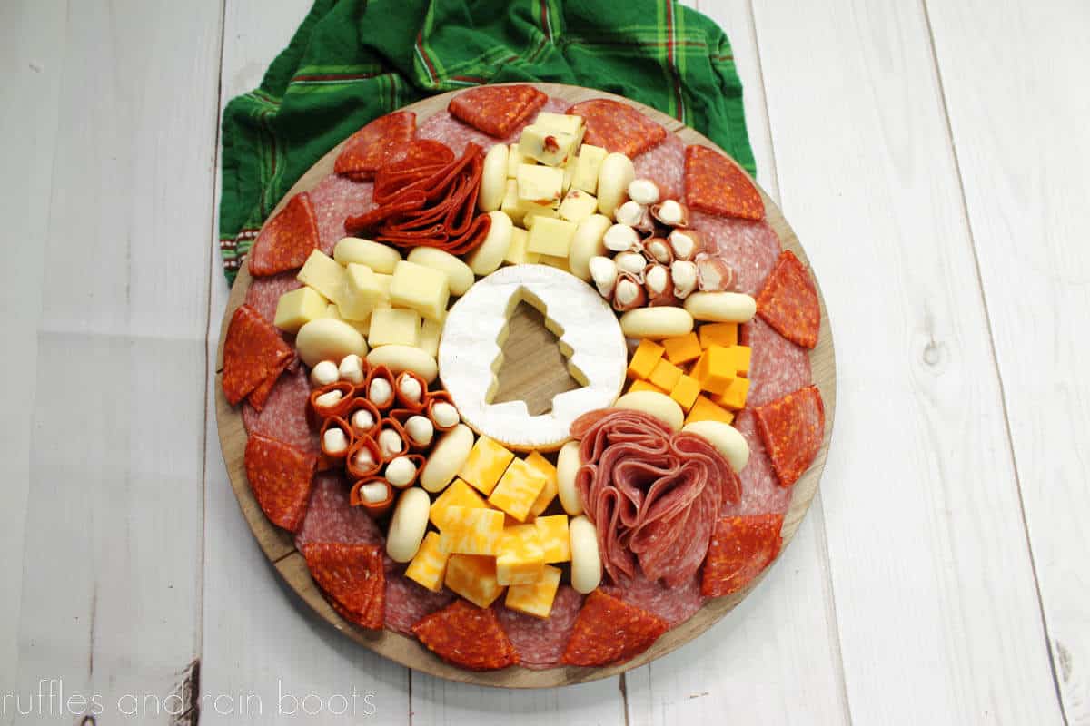 H close up overhead view of a holiday charcuterie board with a variety of meats and cheese with a green plaid napkin on a white wood surface.