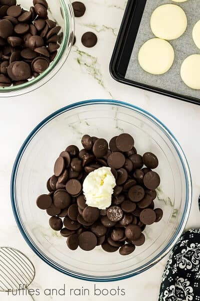 Vertical image of a large glass bowl with vegetable shirtening and chocolate candy melts, next to a smaller bowl of chocolate candy melts and a pan of a few flattened peppermint patties next to black and white fabric on a white marble surface.