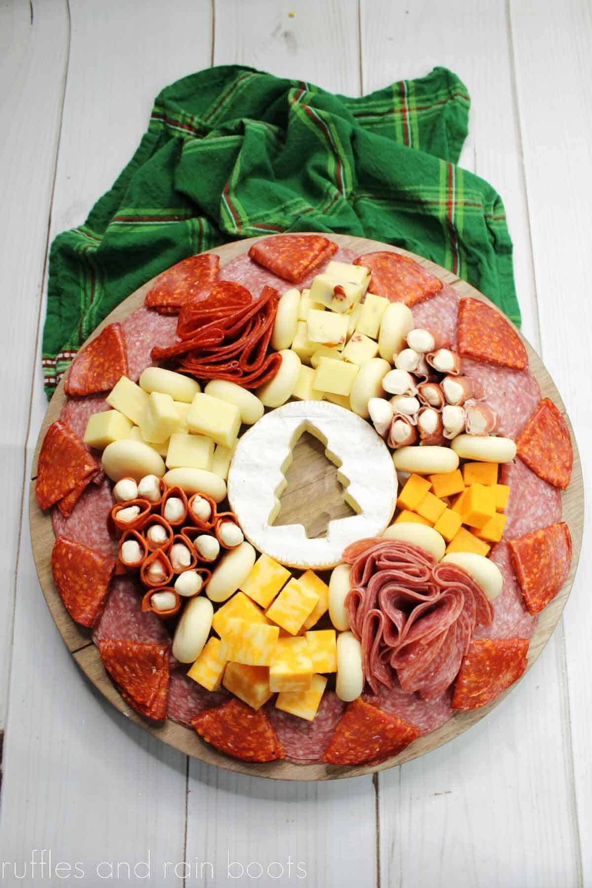 V Overhead view of a holiday charcuterie board with a variety of meats and cheese with a green plaid napkin on a white wood surface..