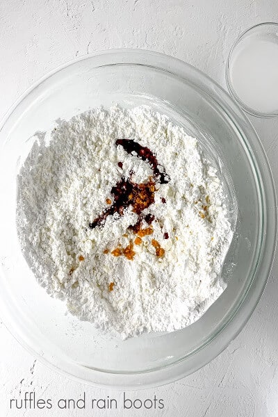 Vertical image of a bowl of powdered sugar, blood orange syrup, and orange peel zest, next to a cup of heavy whipping cream on a white surface.