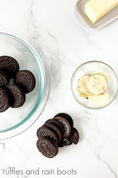 V Overhead image of several Oreo cookies in a round glass bowl, next to a stick of butter, next to a small round glass bowl of the Oreo filling and several opened Oreo cookies on a marble surface.