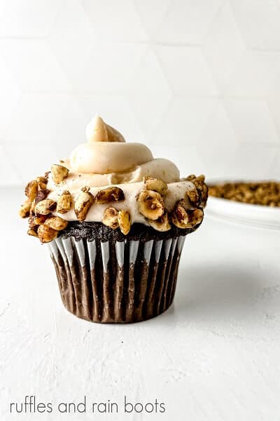 Vertical close up of a frosted cupcake with hazelnuts around the bottom of the frosting on a white surface.