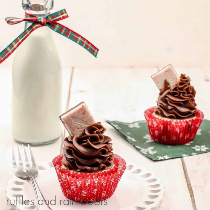 V 2 peppermint cupcakes with mocha frosting and peppermint candies, one on a white plate and ne on a green napkin, next to a bottle of milk with a ribbon tied around the neck on a white and red stripe towel on a wood surface