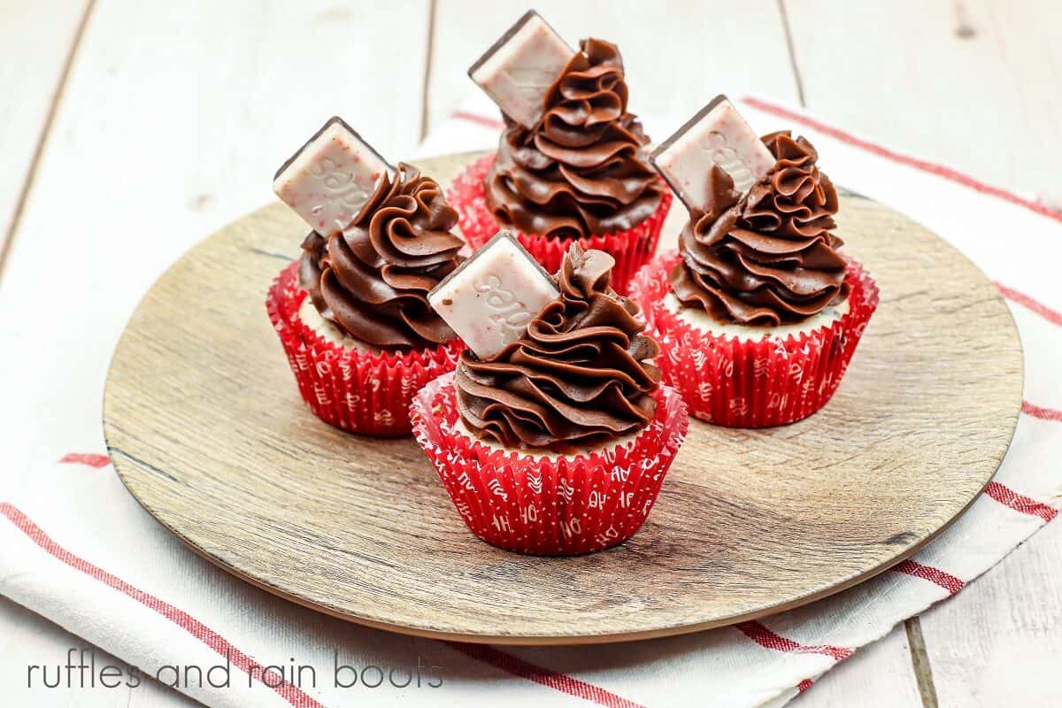 H 4 cupcakes with chocolate frosting and a peppermint candy on top of a wood grain plate on a white and red striped towel on a wood surface