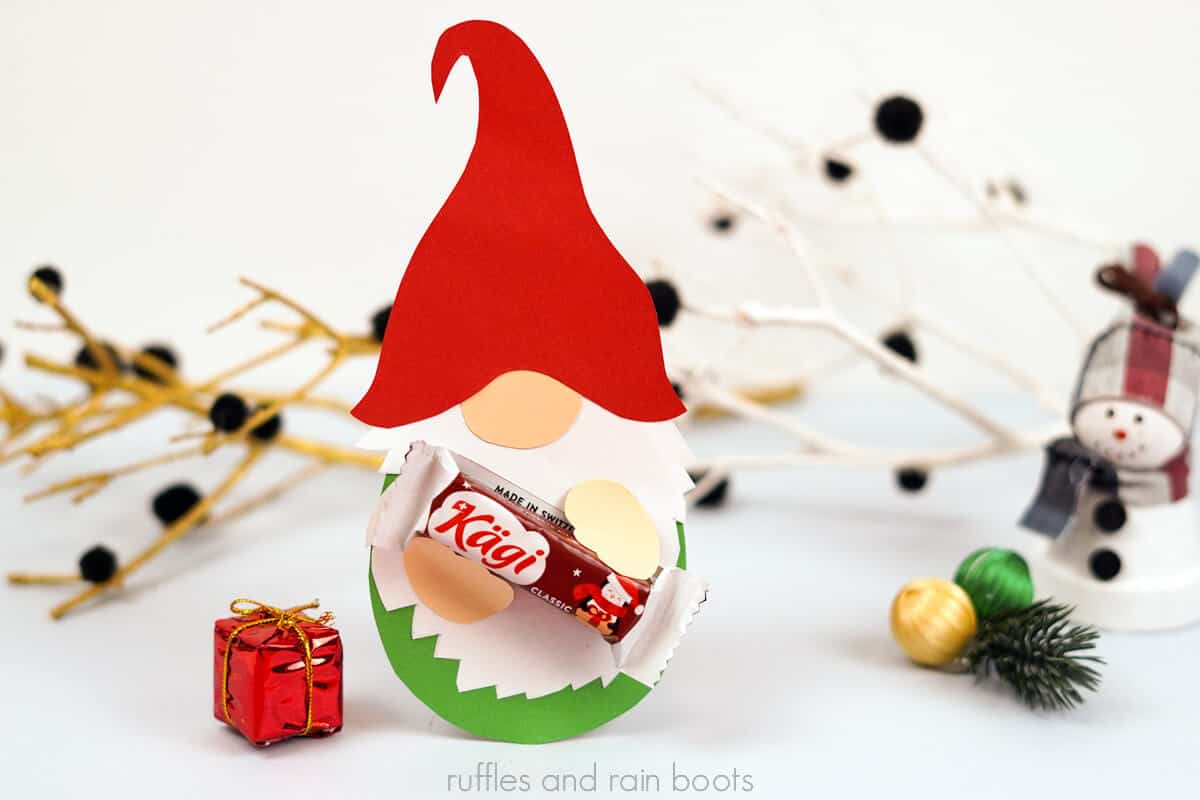 Horizontal image of a red hat gnome holding a piece of chocolate in front of a holiday themed background.