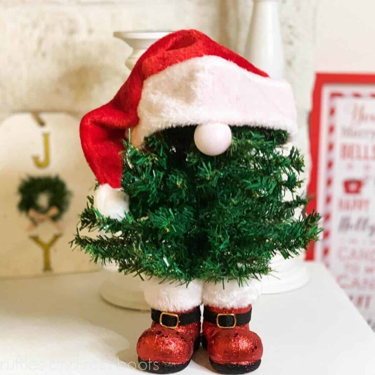 Standing Christmas gnome made with faux trees, a boot ornament, and a red Santa hat in front of a holiday background.