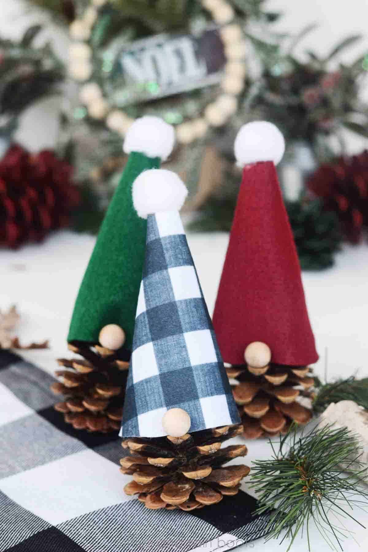 Vertically oriented image showing three gnomes made from felt hats and pinecones with wood bead noses on holiday background.