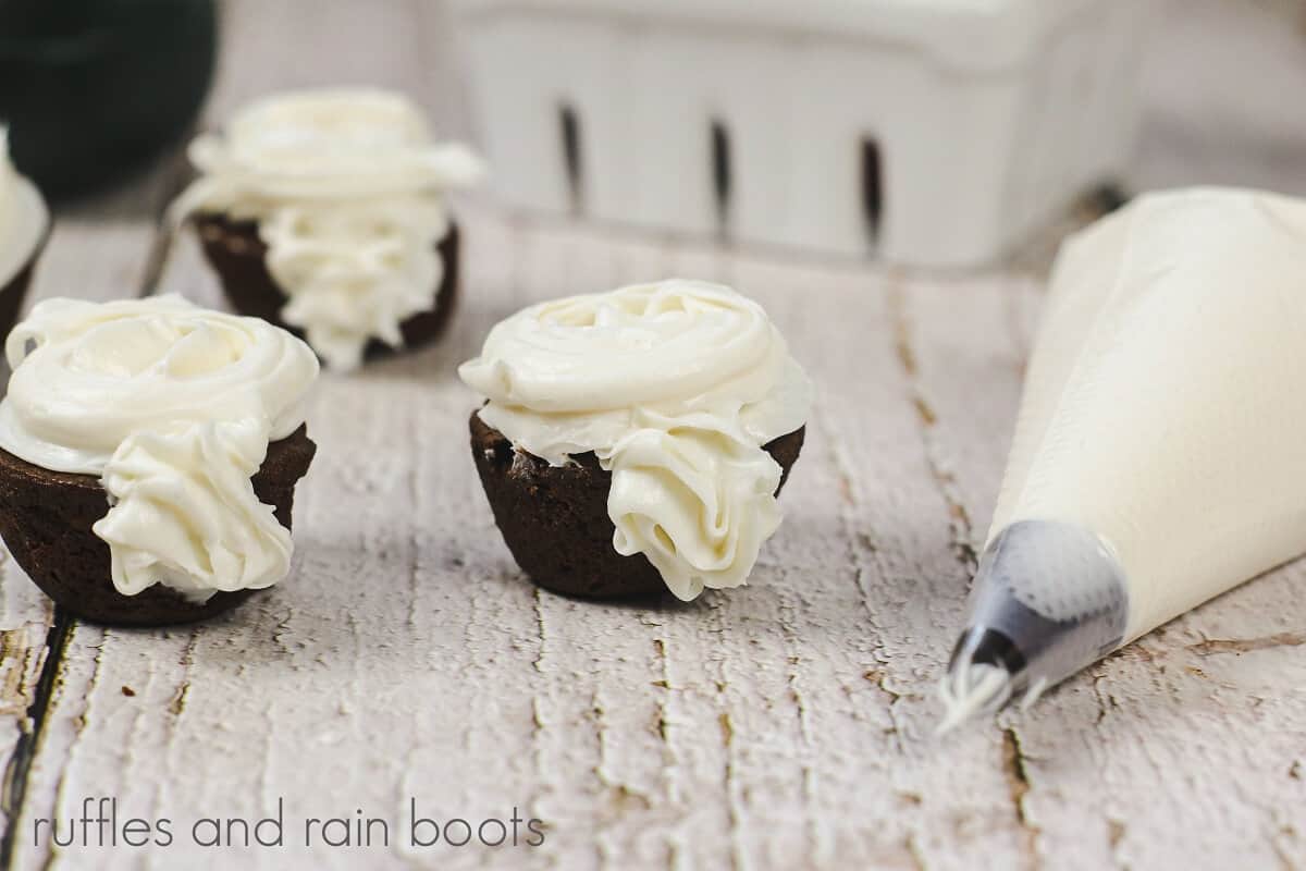 V 3 brownie bites next with vanilla frosting on top next to a piping bag with a white berry basket in the background on a wood surface.