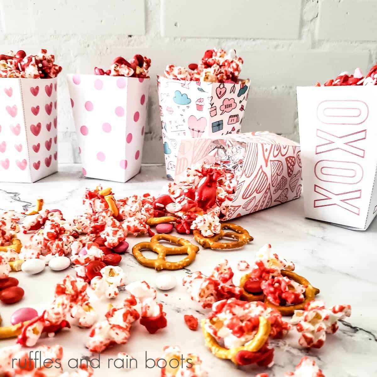 Close up shot of 5 white vertical boxes, one laying down, printed with various Valentine's patterns, filled with popcorn mix with some of the mix scattered on a marble surface.