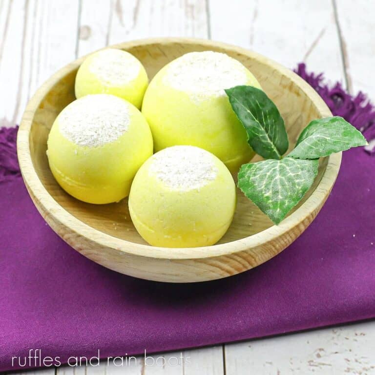 A wooden bowl filled with 4 lemon exfoliating bath bombs with faux leaves on a purple towel on a weathered white wood surface.
