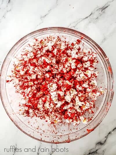 Round glass bowl with popcorn covered in melted red chocolate on a marble surface.