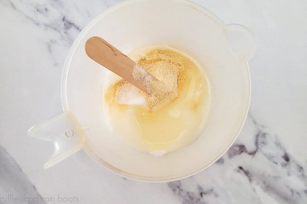 Overhead image of melted white melt and pour soap, sweet almond oil and powdered honey and a wooden craft stick in a plastic measuring cup on a white marble surface.