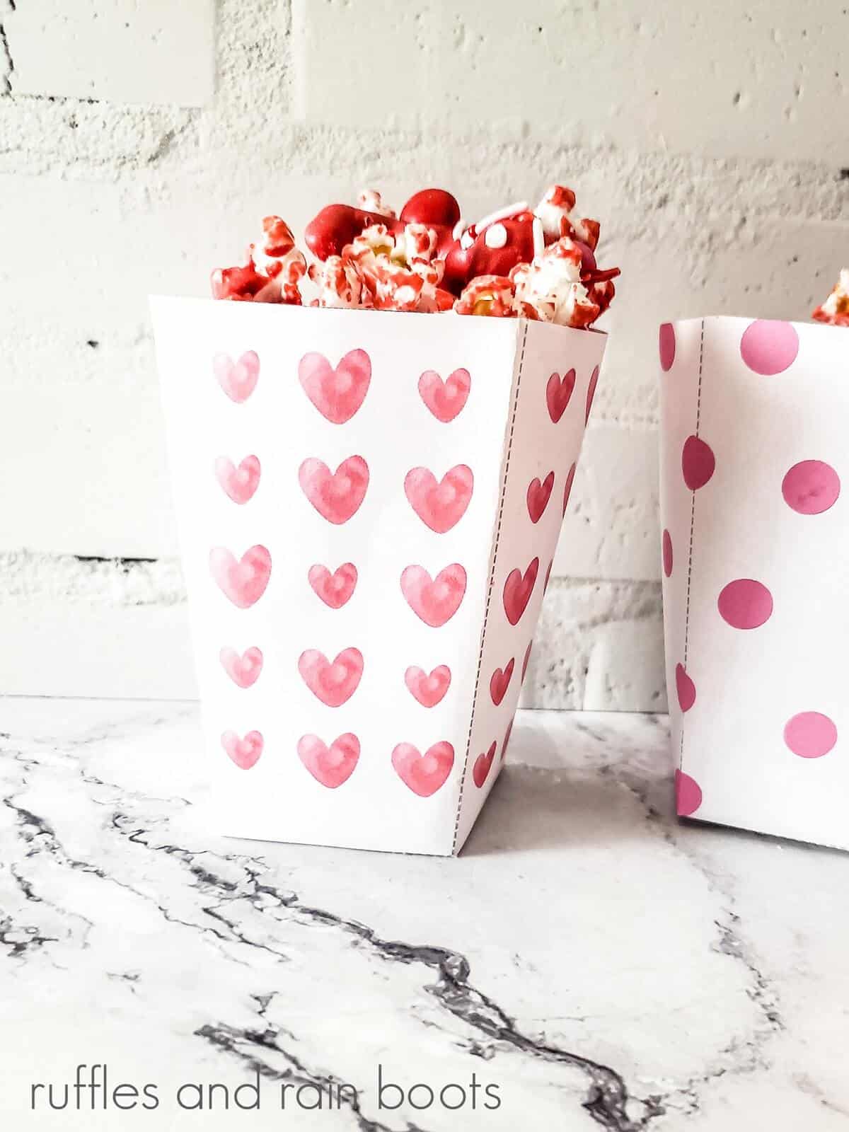 2 white vertical boxes, one with red hearts and one with red circles filled with popcorn mix on a marble surface.