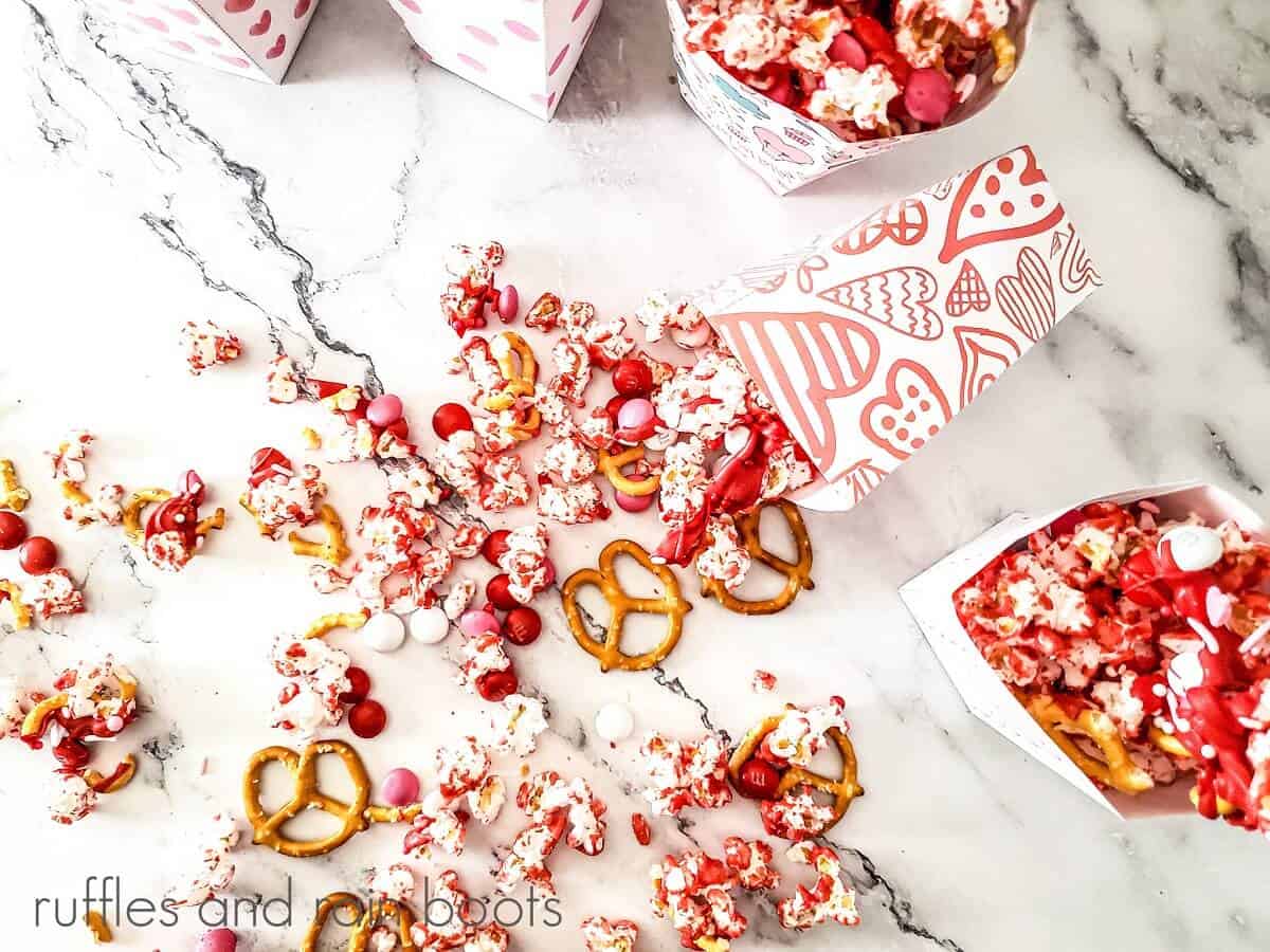 5 white vertical boxes, one laying down, printed with various Valentine's patterns, filled with popcorn mix with some of the mix scattered on a marble surface.