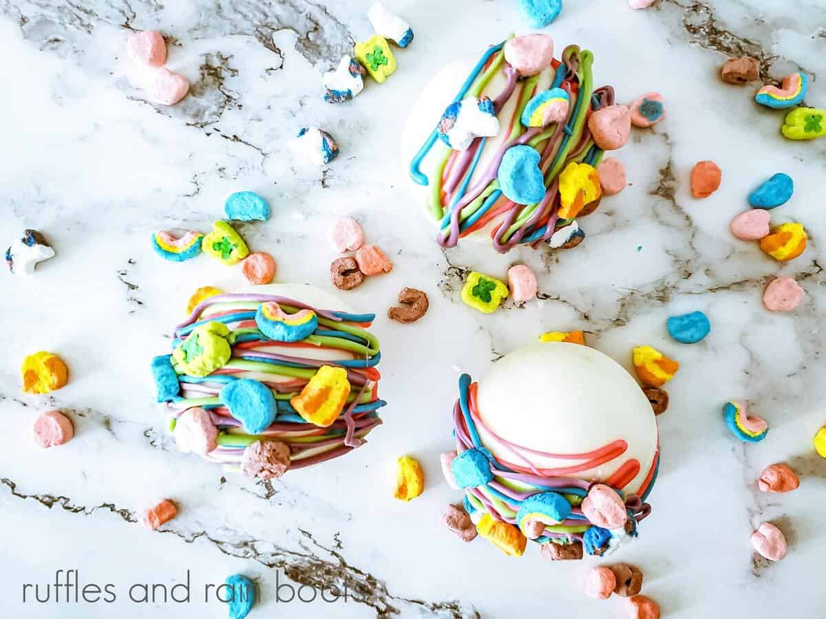 Overhead photo of 3 Lucky Charms Hot Cocoa Bombs on a white marble surface with scattered marshmallows.