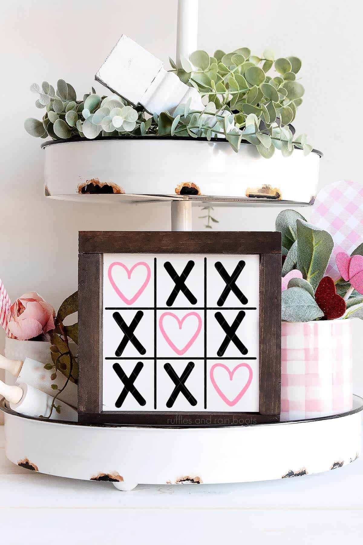 Vertical image of a heart tic tac toe SVG in pink and black vinyl on white dollar store sign placed on decorated tiered tray.