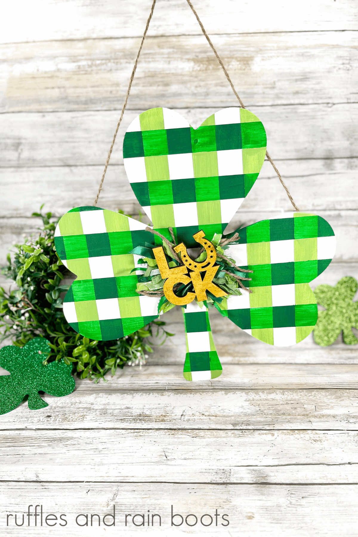 Buffalo Plaid Wooden Shamrock hanging from a piece of twine on a white weathered wood background with faux greenery and shamrocks in the background,