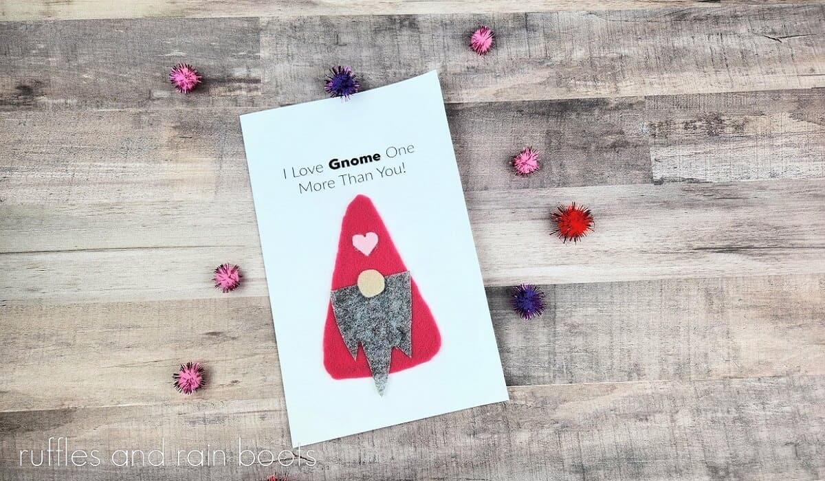 Easy Gnome Valentine's Day card on a weathered wood surface surrounded by colorful pom poms,