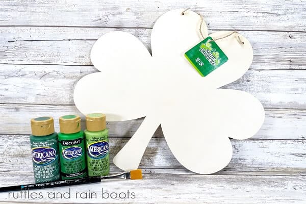 Supplies for the Plaid Wooden Shamrock Sign including the wooden shamrock, a variety of green craft paint and a paintbrush, on a white weathered wood background.