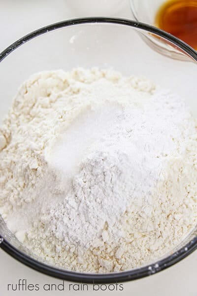 Large round glass bowl with flour and other dry ingredients next to a small bowl of vanilla extract on a white surface