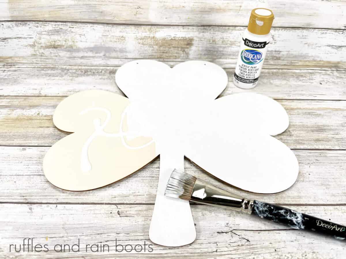 Wooden shamrock, painted white, with a bottle of white craft paint and a paintbrush, on a white weathered wood background.