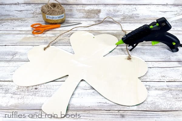 White wooden shamrock with a hot glue gun, scissors and a ball of twine in the background on a white weathered wood background.