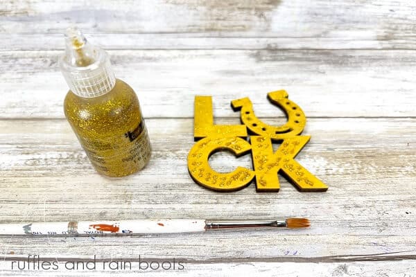 Wooden LUCK sign with a bottle of gold glitter paint and a paintbrush on a white weathered wooden background.