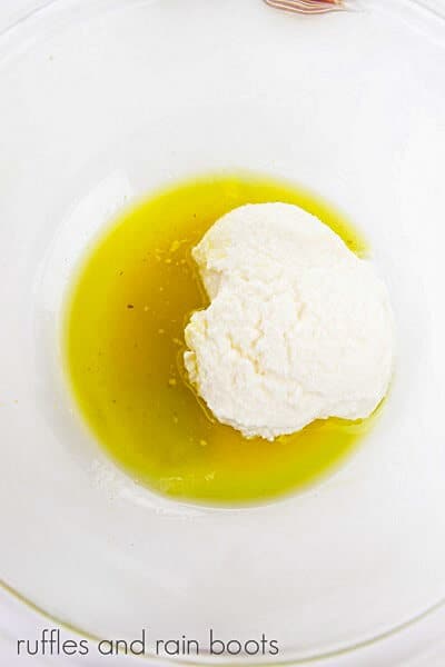 Overhead image of a round glass bowl with olive oil and ricotta cheese on a white surface.