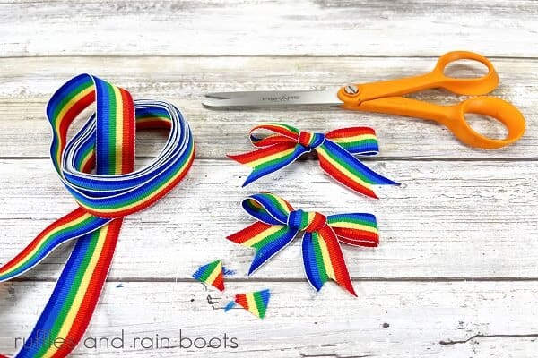 A pair of scissors and rainbow ribbon with 2 bows on a distrssed white wood background