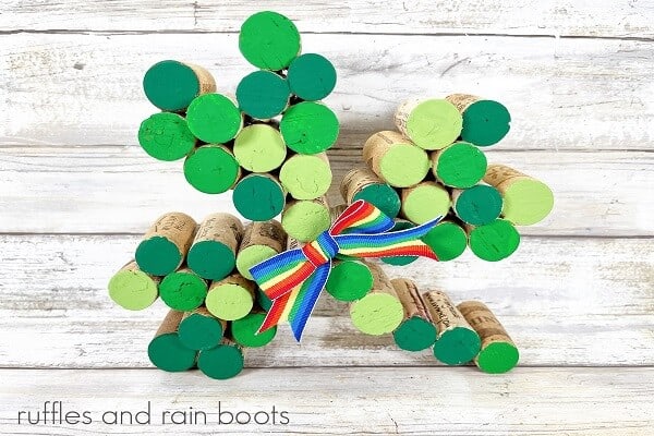 Close up photo of painted wine cork shamrock with a rainbow bow on a distressed white background