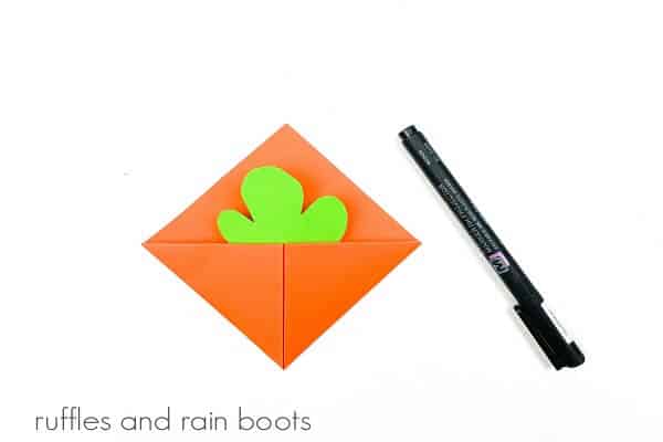 An orange folded paper bookmark with a green leaf cut-out next to a black marker on a white surface.