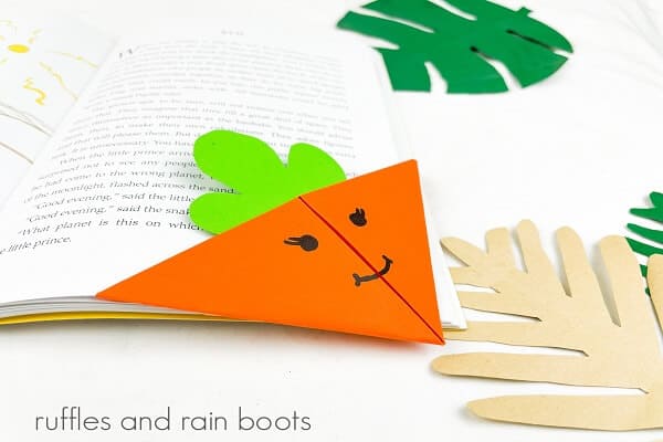 Close up of an orange origami paper carrot bookmark on the bottom right hand corner of a book with faux khaki and green palm leaves on a white surface.