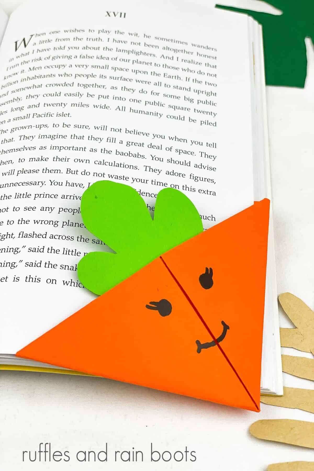 A close up of an orange carrot bookmark on the bottom right hand corner of a book, next to a khaki and green faux palm leaf on a white surface.