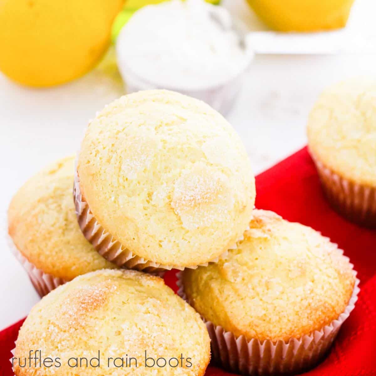 A batch of Homemade Lemon Ricotta Muffins on a red napkin with lemons in the background on a white surface.