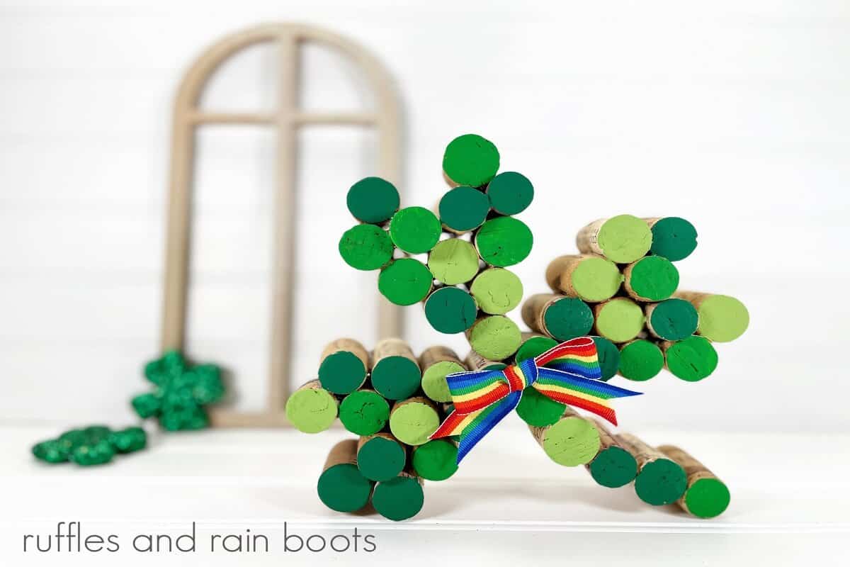 A close up photo of a painted wine cork shamrock in various shades of green, with a rainbow bow with a wooden window and St. Patty’s Day decorations on a white weathered wood background.