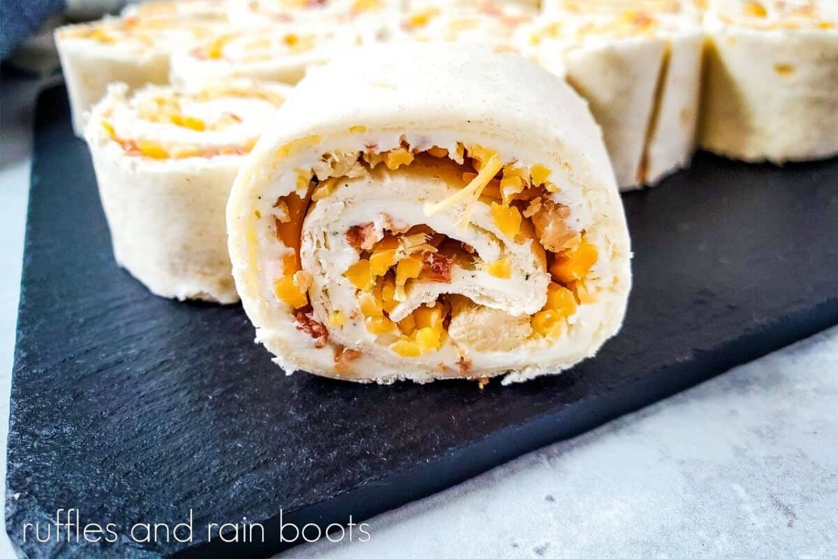 Close up image of a crack chicken pinwheel sandwich on a black marble slab, next to a small bowl of shredded chicken and a grey and white striped towel on a white marble surface.